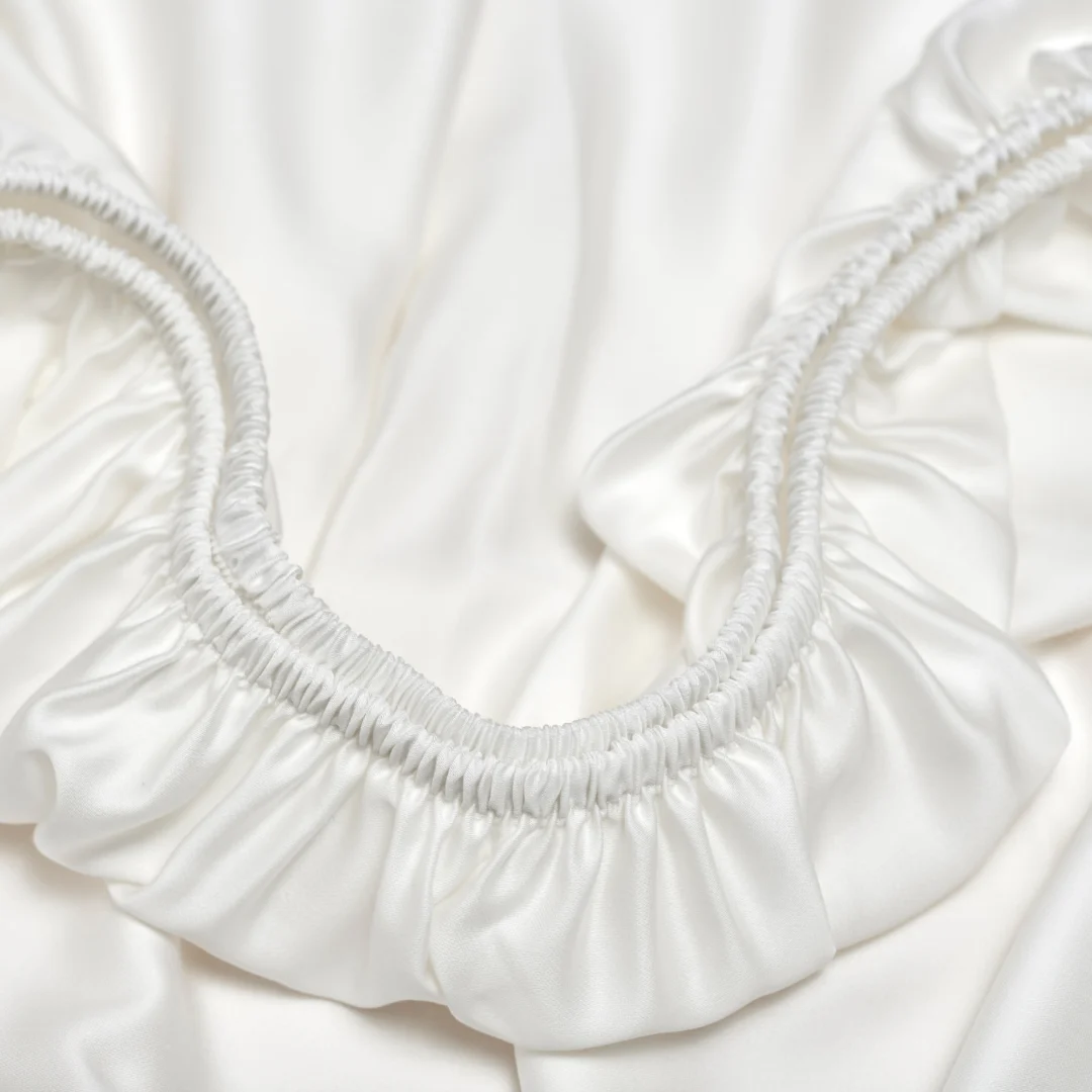 Close-up van een wit hoeslaken met elastiek, zachte glans en subtiele plooien. Het materiaal oogt glad en luxe. Zachte belichting.