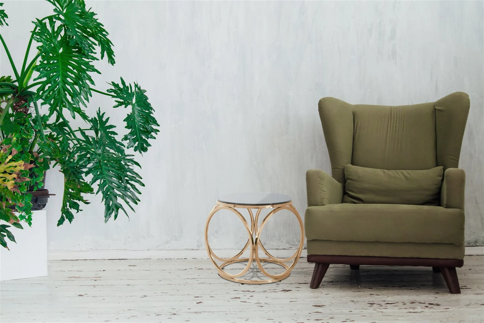Een groene fauteuil met kussen staat naast de Bijzettafel Grijs Staal Chique, een chique grijs stalen bijzettafel. Links staat een grote, bebladerde potplant tegen een witgeschilderde muur en een verweerde houten vloer.