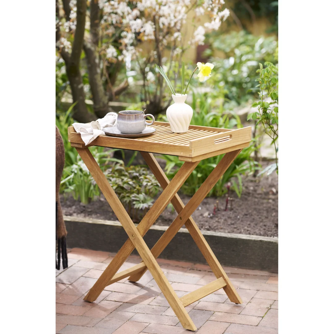 De Ronda Dienbladtafel, een compacte houten klaptafel, staat op een stenen patio in een tuin met een witte vaas met gele bloemen, een keramische mok en een gevouwen kleed, omringd door weelderig groen en bloeiende planten.