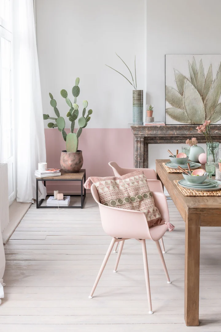 Een moderne eetkamer met de Bijzettafel Zwart, Bruin (60x41 CM), een vierkante metalen bijzettafel, in combinatie met roze stoelen, pastelkleurige decoraties, een grote cactus en een open haard versierd met planten en kunst; natuurlijk licht vult de ruimte.