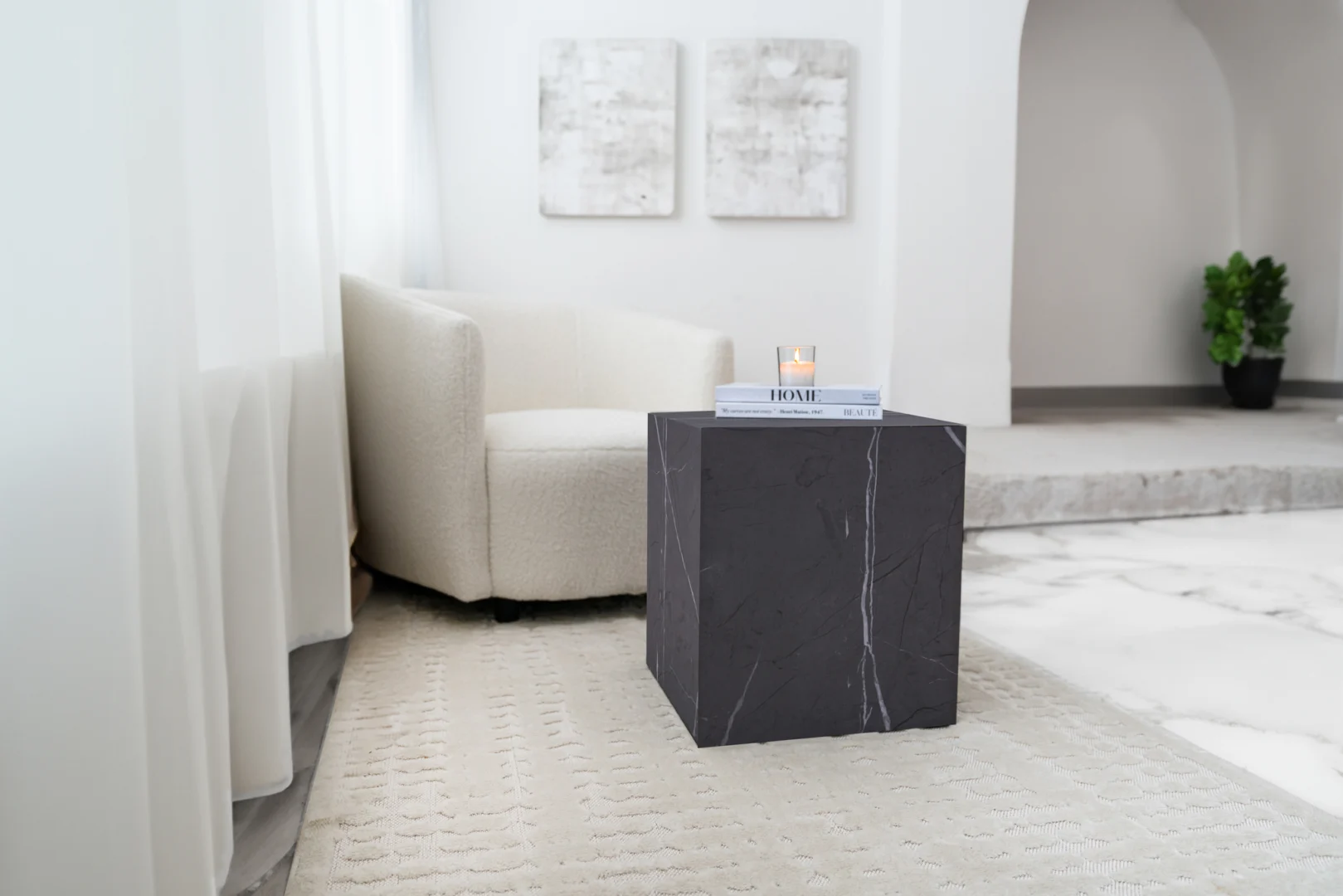 Een moderne woonkamer met een crèmekleurige fauteuil, een Bijzettafel Grijs Mdf Design met een kaars en boeken, een vloerkleed met structuur, abstracte muurkunst en een plant in pot op de achtergrond.