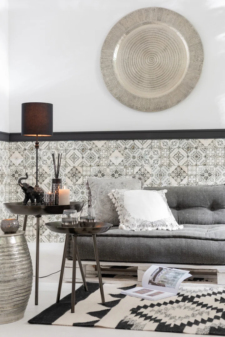 Een moderne woonkamer met een grijze bank, decoratieve kussens, gedessineerde wandtegels, zwarte tafellampen, kaarsen, een olifantenbeeld, een ronde metalen Bijzettafel Donkergrijs Greige 52 x 58 cm en een zwart-wit geometrisch vloerkleed.