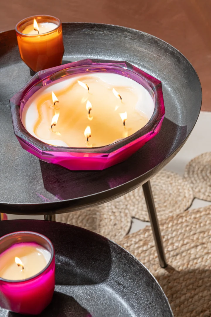 Een roze glazen schaalkaars met verschillende brandende lonten staat op een Bijzettafel Donkergrijs, Greige 52 x 58 CM Rond Aluminium, vergezeld van twee kleine oranje kaarsen; een geweven mat is gedeeltelijk zichtbaar op de vloer.