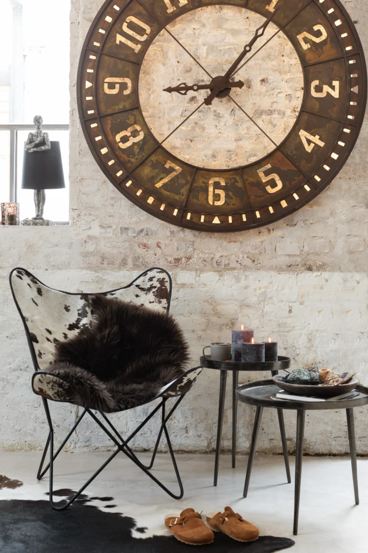 Een gezellig hoekje met een vlinderstoel van koeienhuid, een bontworp, een ronde aluminium tafel van Bijzettafel Donkergrijs Greige 61x61 cm met kaarsen en hapjes, bruine klompen op een tapijt van koeienhuid en een grote rustieke wandklok op een muur van witte bakstenen.