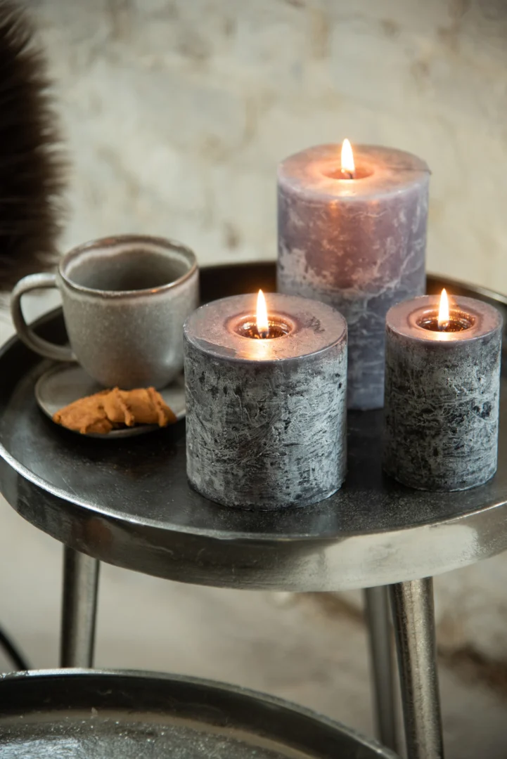 Drie grijze pilaarkaarsen worden aangestoken op de Bijzettafel Donkergrijs, Greige 61 x 61 cm ronde aluminium tafel, naast een grijze keramische beker en een bord koekjes, waardoor een gezellige en warme sfeer ontstaat.