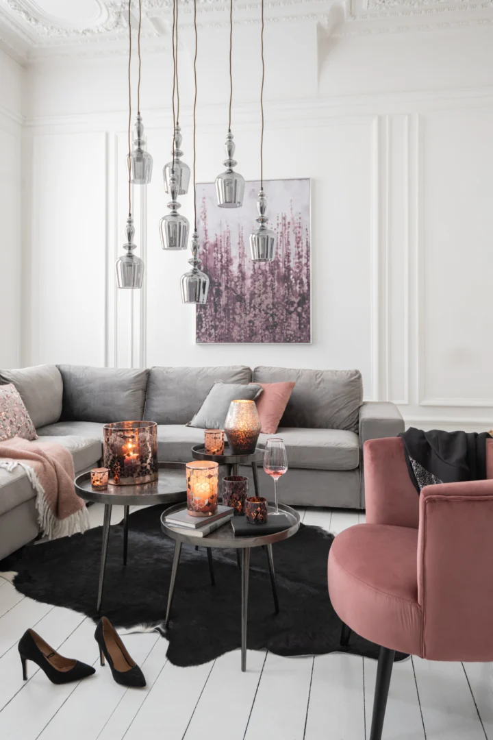 Een moderne woonkamer heeft een grijze bank, een roze accentstoel en hanglampen. Twee Bijzettafel Donkergrijs, Greige 61 x 61 CM ronde aluminium tafels tonen kaarsen en een wijnglas. Roze kunst hangt boven zwarte hakken op witte houten vloeren.