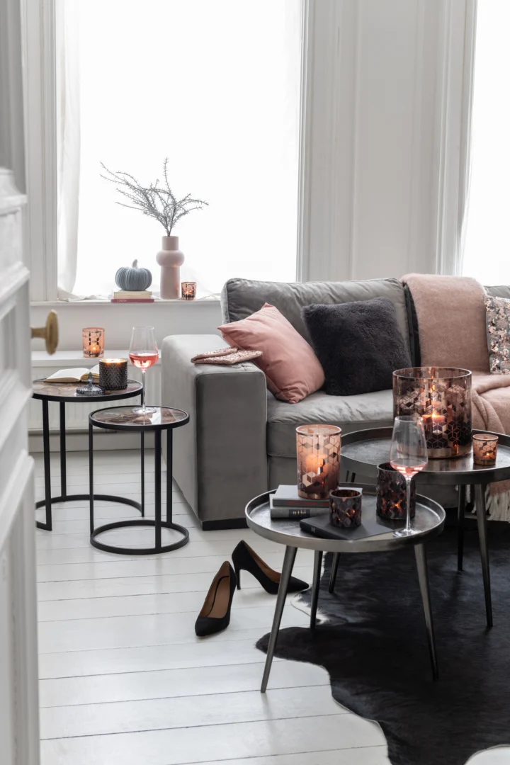 Een moderne woonkamer met een grijze bank, roze en zwarte kussens, de Bijzettafel Donkergrijs, Greige 61 x 61 cm rond aluminium bijzettafel met kaarsen, glazen, boeken, een roze vaas met takken en zacht natuurlijk licht van grote ramen.