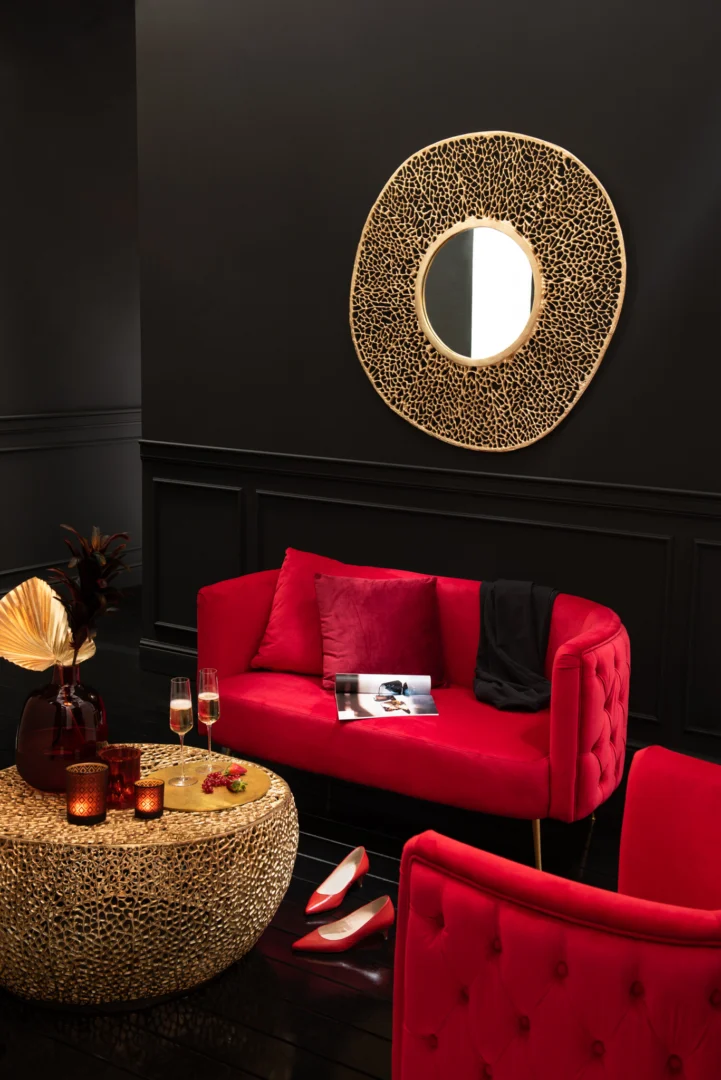 Een stijlvolle woonkamer met een dieprode fluwelen bank, gouden accentkussens en de Salontafel Goud 85 x 85 CM Rond Aluminium salontafel. Een ronde goudomrande spiegel siert een donkere muur. Rode hakken en champagneglazen voegen luxe toe.
