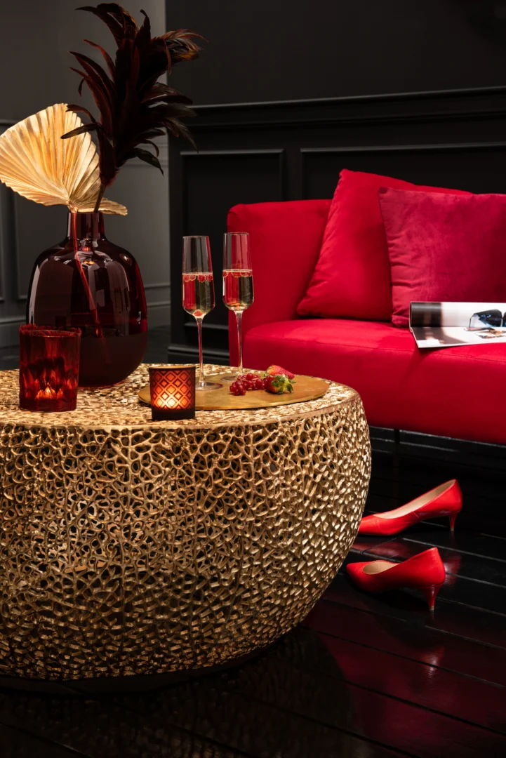 Een stijlvolle woonkamer met de Salontafel Goud 32 CM Rond Aluminium, twee champagneglazen, kaarsen, decoratieve vazen, een felrode bank met kussens, een tijdschrift en rode hoge hakken op een donkere houten vloer.