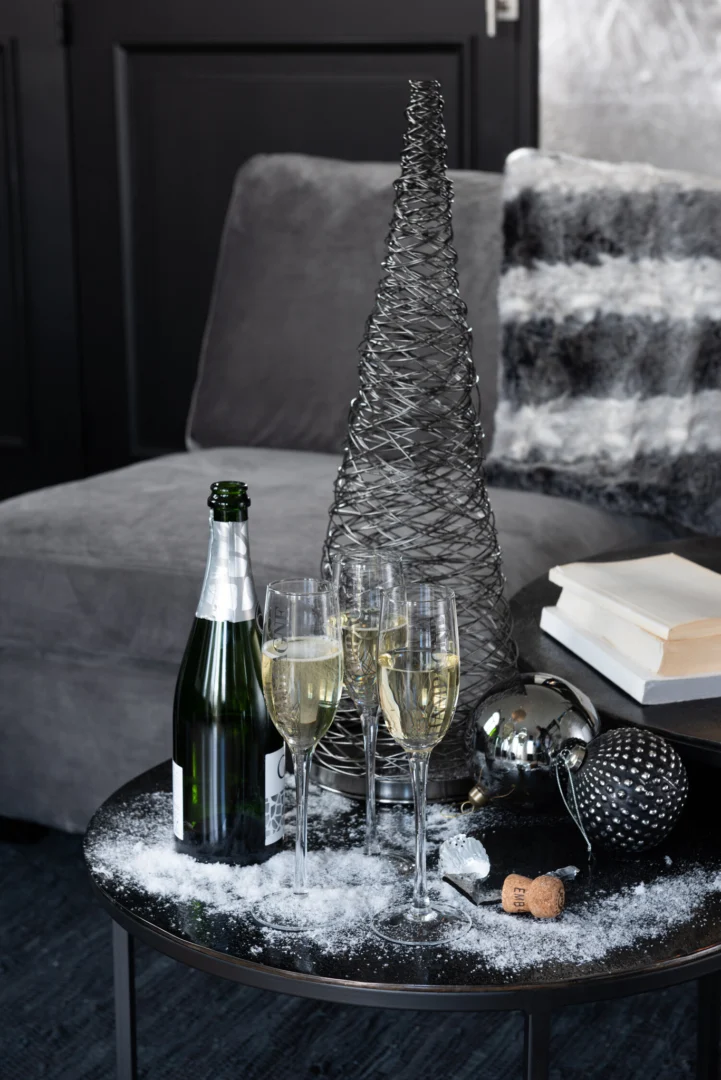 Op de Bijzettafel Zwart Rond Aluminium staan drie champagneglazen, een fles met kurk, feestelijke ornamenten, boeken, nepsneeuw en een kerstboom van zilverdraad, met een grijze bank en bontkussen op de achtergrond.