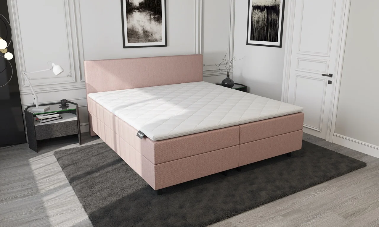 Boxspring oud roze met opbergruimte, gezien in een lichte kamer. Het bed heeft een zachtroze kleur en een wit matras met ruitpatroon.