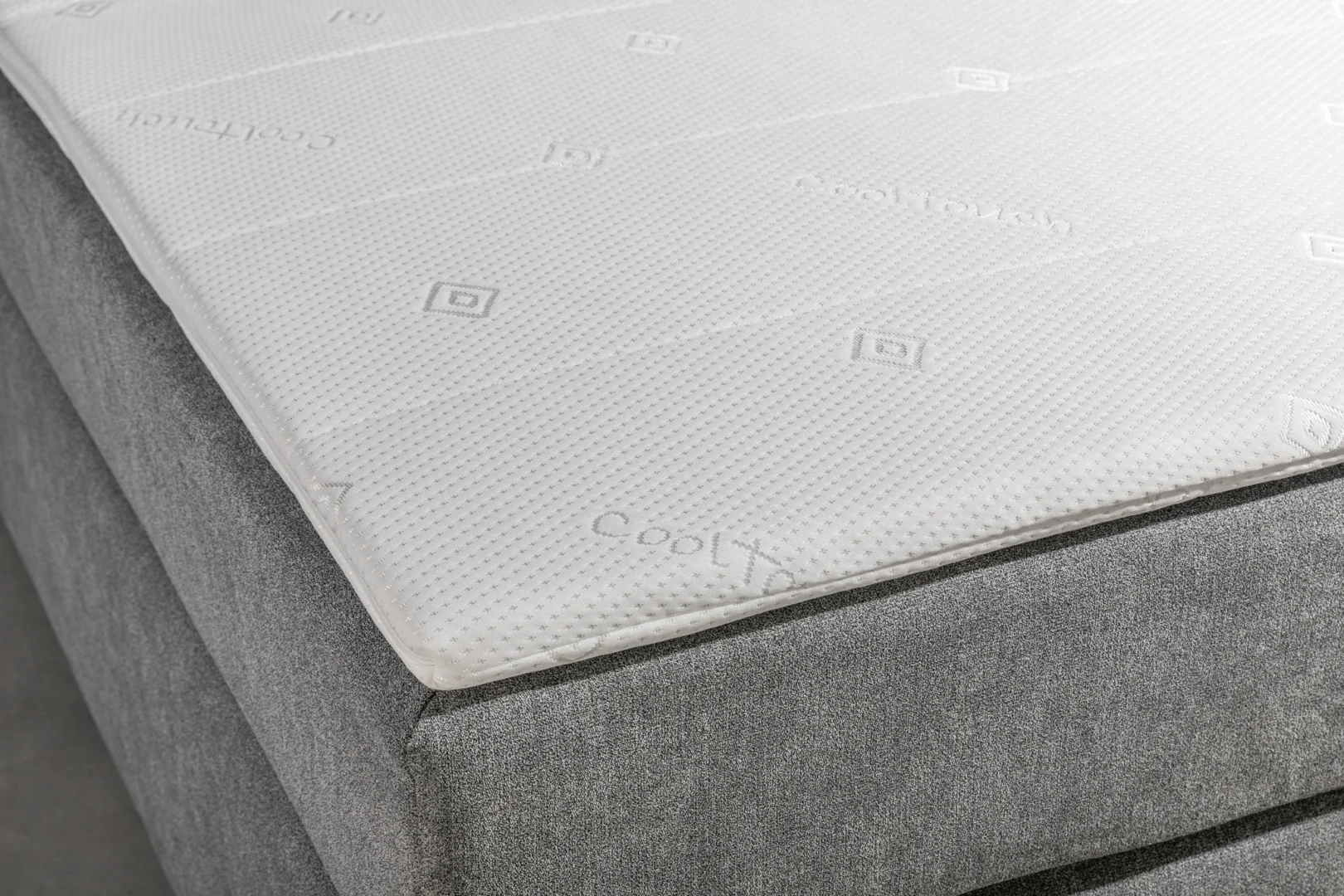Detailopname van een boxspring met een witte topper. Zachte belichting en een grijze achtergrond. Textuur van de topper is goed zichtbaar.