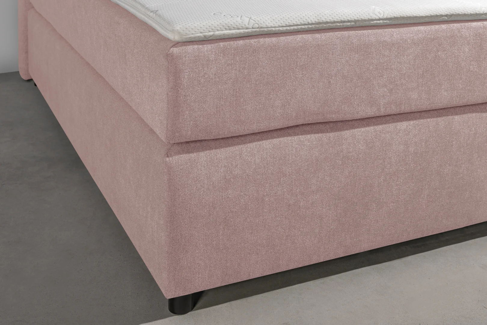 Detailopname van een roze stoffen boxspring met een wit matras. De boxspring heeft zwarte poten en staat op een grijze ondergrond.