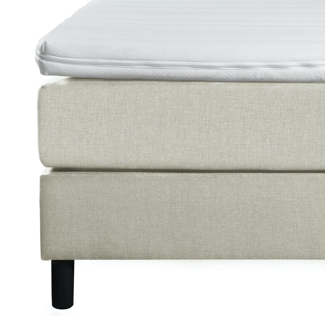 Detailopname van een beige boxspring met een witte koudschuimtopper. Zwarte poten, zachte belichting, textuur van de stof is goed zichtbaar.