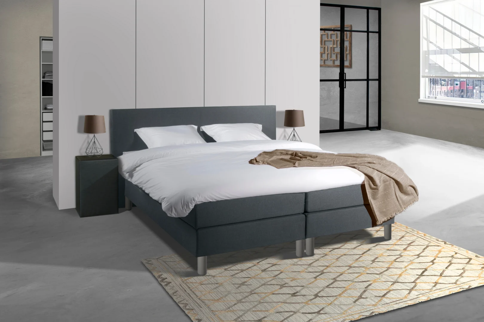 Antraciet houten boxspring met witte lakens en een beige sprei. De boxspring staat op een beige vloerkleed in een lichte kamer.