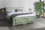 Groene boxspring van stof in een lichte kamer. Het bed heeft wit beddengoed en een grijze plaid. Op de vloer ligt een grijs kleed.