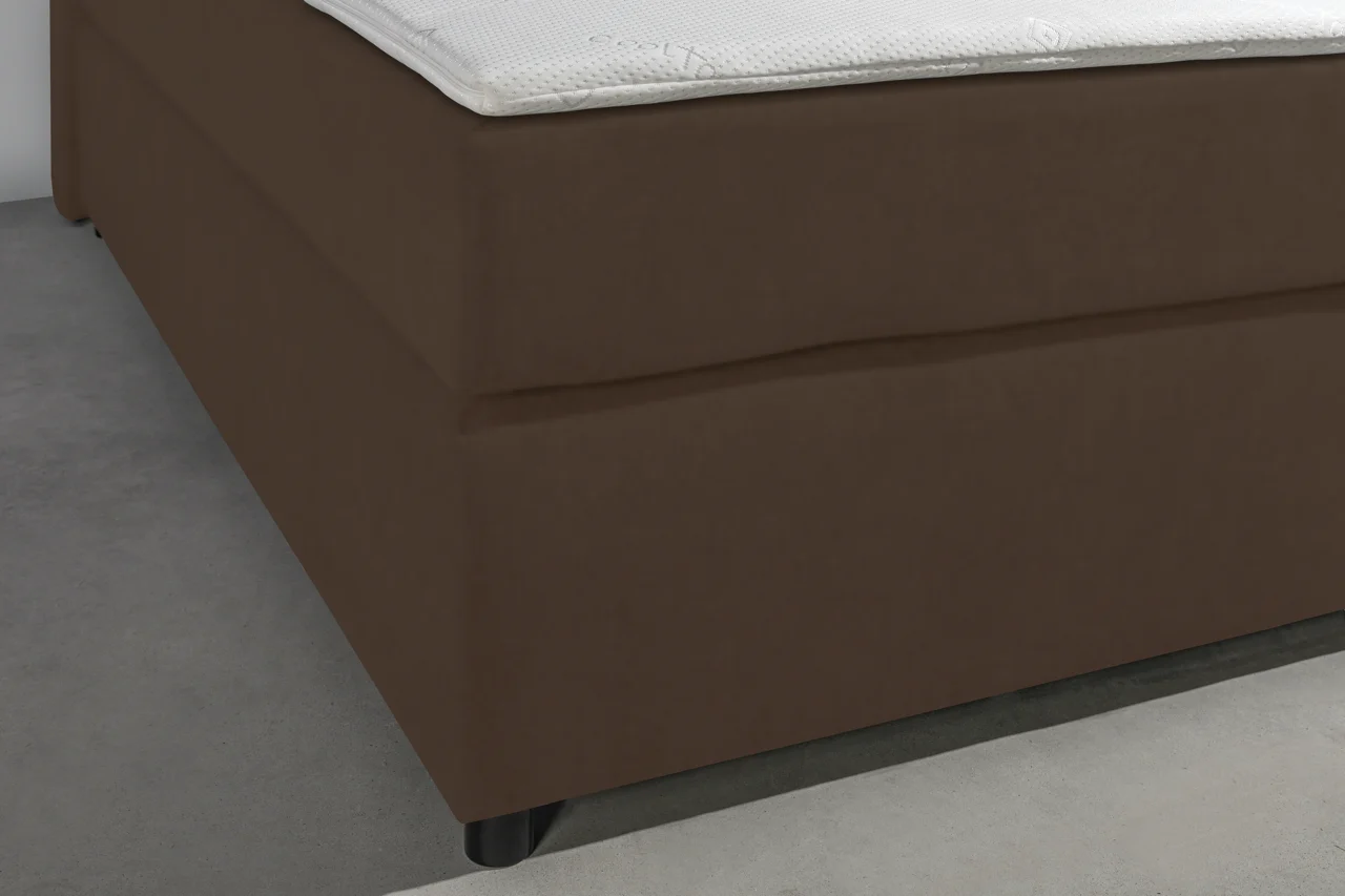 Detailopname van een bruine boxspring met lederlook en een witte topper, lage hoek, zachte belichting op een grijze ondergrond.