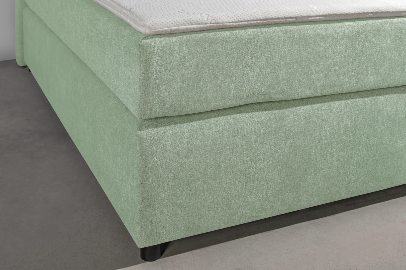 Detailopname van een lichtgroene boxspring met topper. De zachte stof heeft een subtiele textuur. Houten frame en donkere poten zijn zichtbaar.