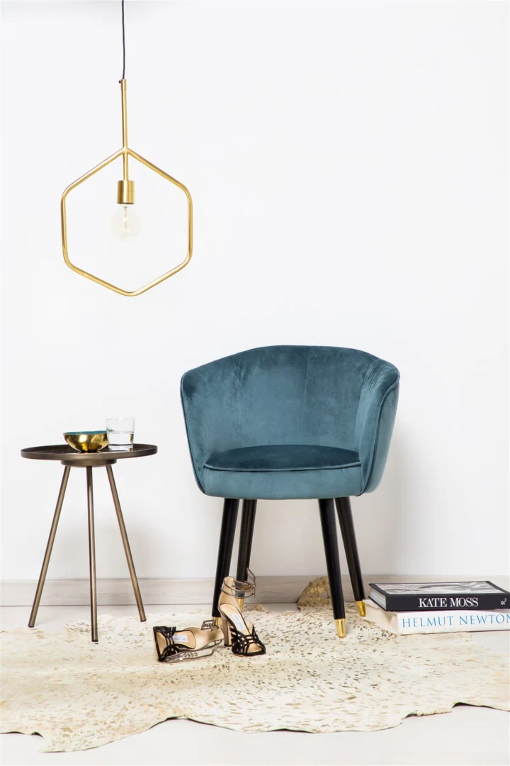 Een modern interieur met een blauw fluwelen stoel, de Bijzettafel Rond Glas met een glas en een schaal, een geometrische hanglamp, schoenen met hakken op een vloerkleed en gestapelde boeken op de vloer bij een witte muur.
