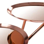 Close-up van twee ronde Bijzettafel Goud, Roze Ijzer Chique dienbladen met koperkleurige spiegeloppervlakken, verbonden door schuine stangen, tegen een witte achtergrond.