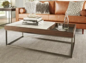 Een moderne woonkamer met een bruine leren bank, een vloerkleed met patroon en de Salontafel Walnoot, Bruin Rvs salontafel met boeken, een waterfles en een glas op een dienblad. De ruimte is gevuld met natuurlijk licht.