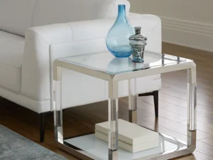 De Jasper bijzettafel in acryl, wit glas en gepolijst roestvrij staal staat naast een witte bank met een blauwe glazen vaas, een decoratieve pot en twee boeken op de onderste plank, op houten vloer met een blauw vloerkleed.