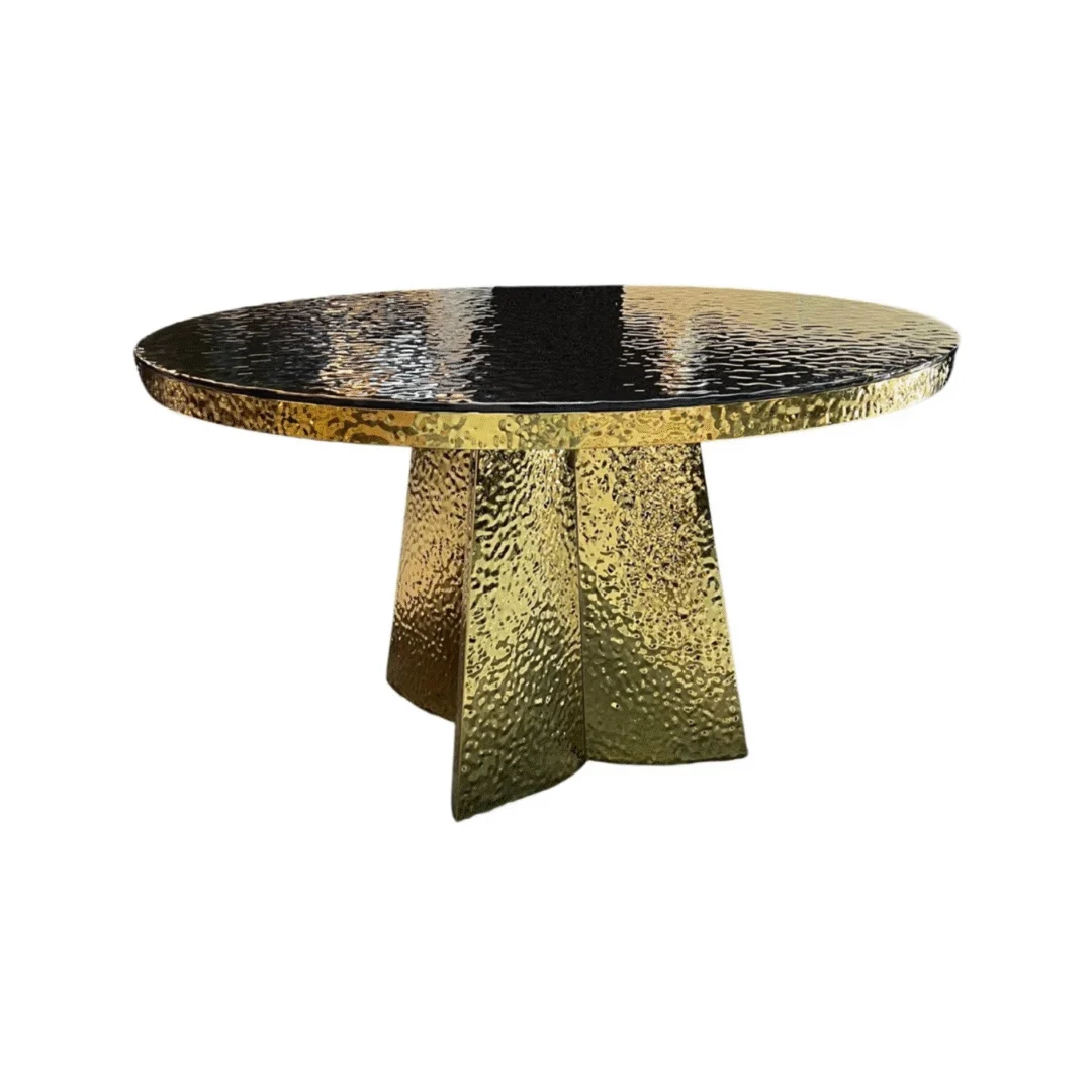 De JIMMY DT GOUD 130 ROND ZWART GESMOLTEN GLAS is een ronde tafel met een glanzend, gehamerd gouden tafelblad en een gestructureerd, reflecterend onderstel, ondersteund door vier brede kruisende poten in het midden.