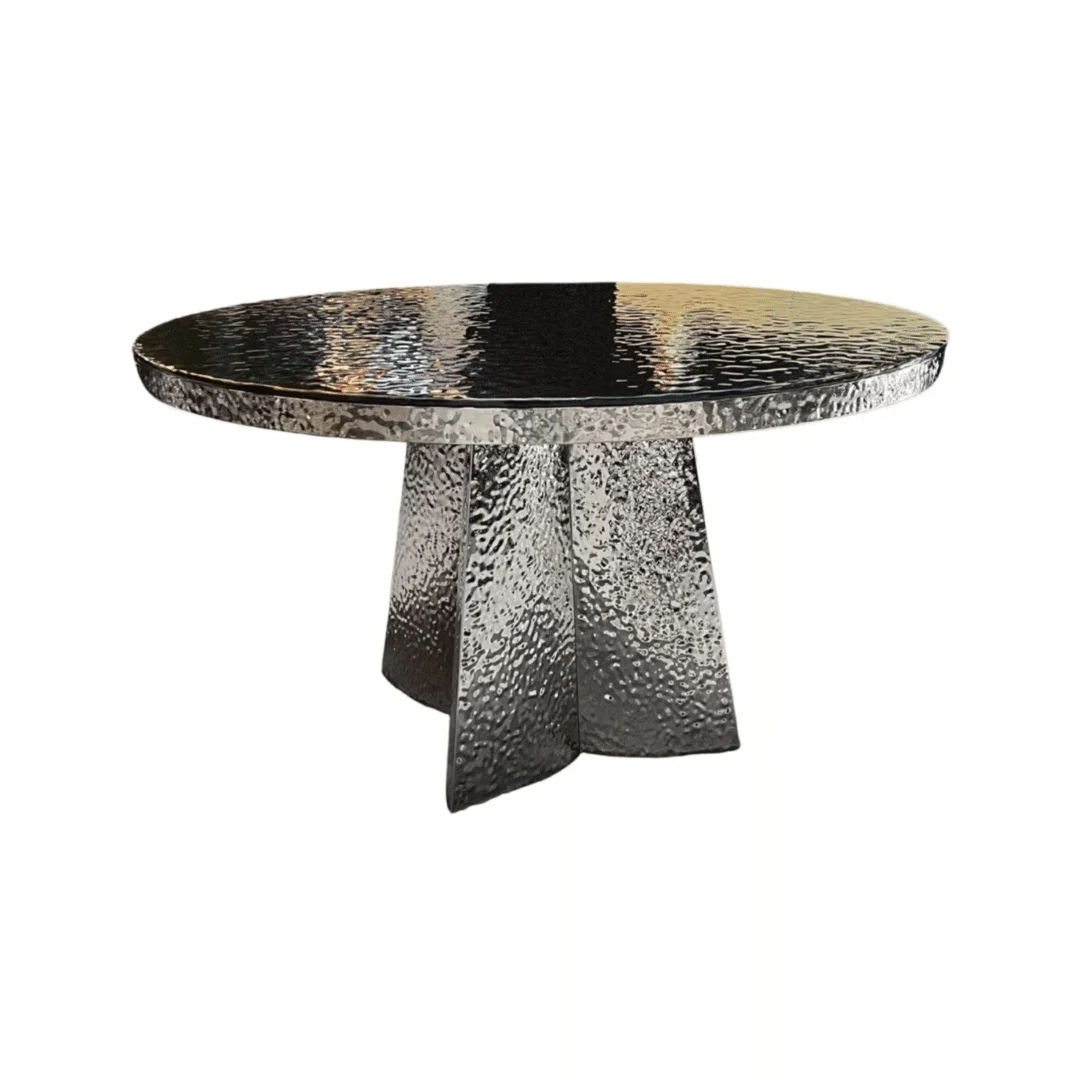 De JIMMY DT ZILVER 130 ROND ZWART GESMOLTEN GLAS is een ronde tafel met een gestructureerd metalen oppervlak en een gehamerd onderstel met drie brede, schuine steunen, tegen een effen witte achtergrond.