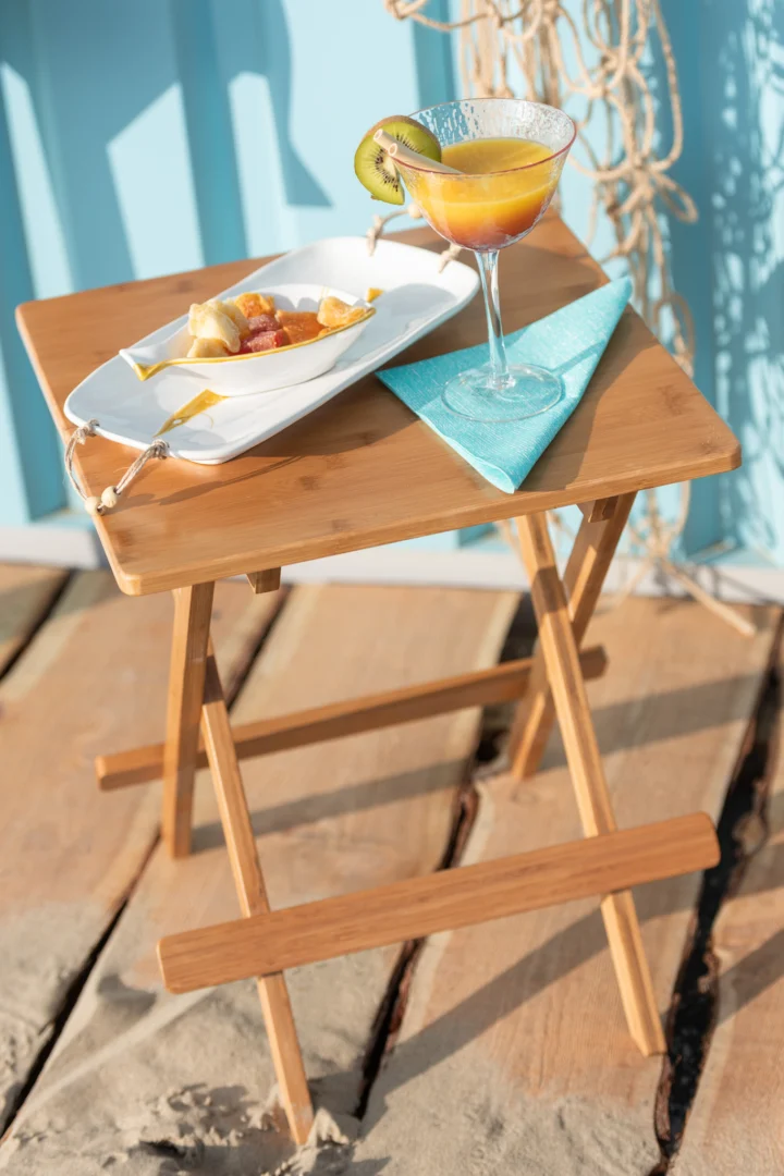 Een Bijzettafel 40 x 48 CM Bamboe houdt een martiniglas met een sinaasappeldrankje en een schijfje kiwi vast, naast een rechthoekig bord met fruitspiesjes en een gevouwen blauw servet, op een houten vlonder.