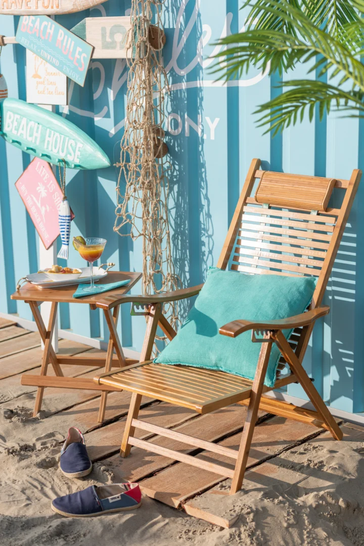 Een houten strandstoel met een groenblauw kussen rust op zanderige planken naast de Bijzettafel 40 x 48 CM Bamboe met drankjes, omringd door nautische tekens, een palm en slip-on schoenen, allemaal tegen een blauwe muur met stranddecoratie.