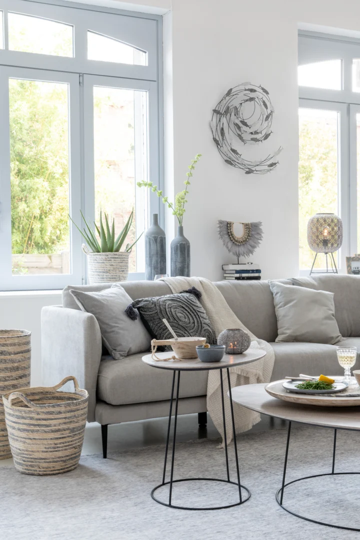 Een moderne woonkamer met een lichtgrijze sectionele bank, kussens met patronen, geweven manden, vazen met groen, grote ramen en de Bijzettafel Greige Zwart 55 CM Ovaal-een strakke, ovale ijzeren bijzettafel met chique decoratiespullen.