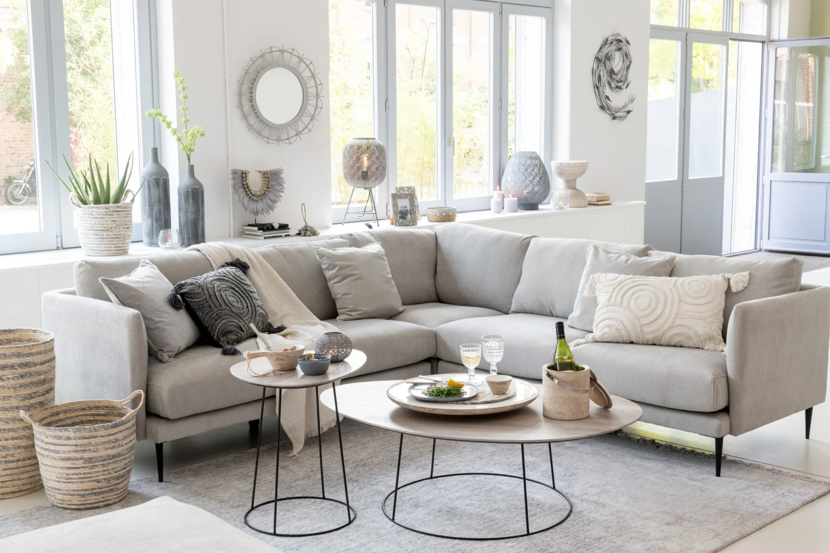 Een moderne woonkamer met een lichtgrijs bankstel, sierkussens, geweven manden, vazen, muurkunst en de Bijzettafel Greige Zwart 55 CM Ovaal ijzeren bijzettafel, allemaal in een lichte ruimte met grote ramen.