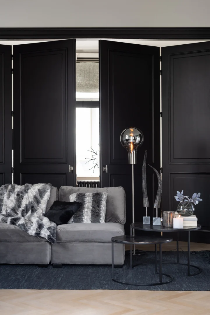 Een moderne woonkamer heeft een grijze bank met gedessineerde dekens, boeken, kaarsen, een glazen globe vloerlamp en de Bijzettafel Zwart 40 CM Rond Aluminium als strakke zwarte geneste salontafels tegen hoge zwarte paneeldeuren.