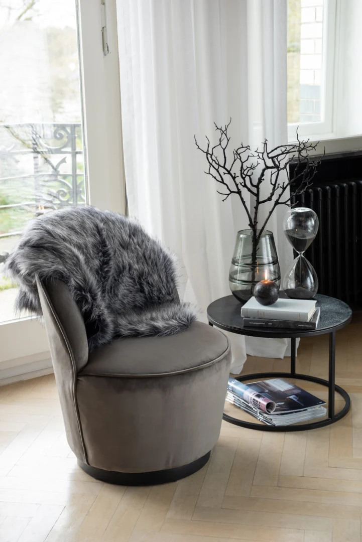 Een gezellige grijze fluwelen stoel met een faux fur throw staat naast de Bijzettafel Zwart 40 CM Rond Aluminium, waarin glazen vazen met kale takken, een zandloper, een kaars en boeken staan. Zonlicht valt door de witte gordijnen bij het raam.