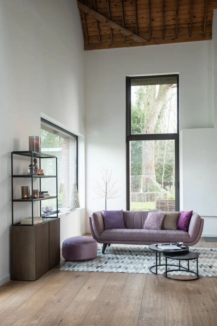 Een moderne woonkamer met hoge plafonds, grote ramen, een paarse bank met kussens, een ronde voetenbank, een geometrisch vloerkleed, een kast tegen witte muren en de Bijzettafel Zwart 40 CM Rond Aluminium salontafel. Natuurlijk licht vult de kamer.