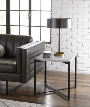In een moderne woonkamer staat de Saxon Bijzettafel in Carrara Marmer en Mat Zwart als bijzettafel, in combinatie met een zwart leren bank, grijze en gouden lamp, klein marmeren kastje, witte bakstenen muur en grote ramen met zwart frame.