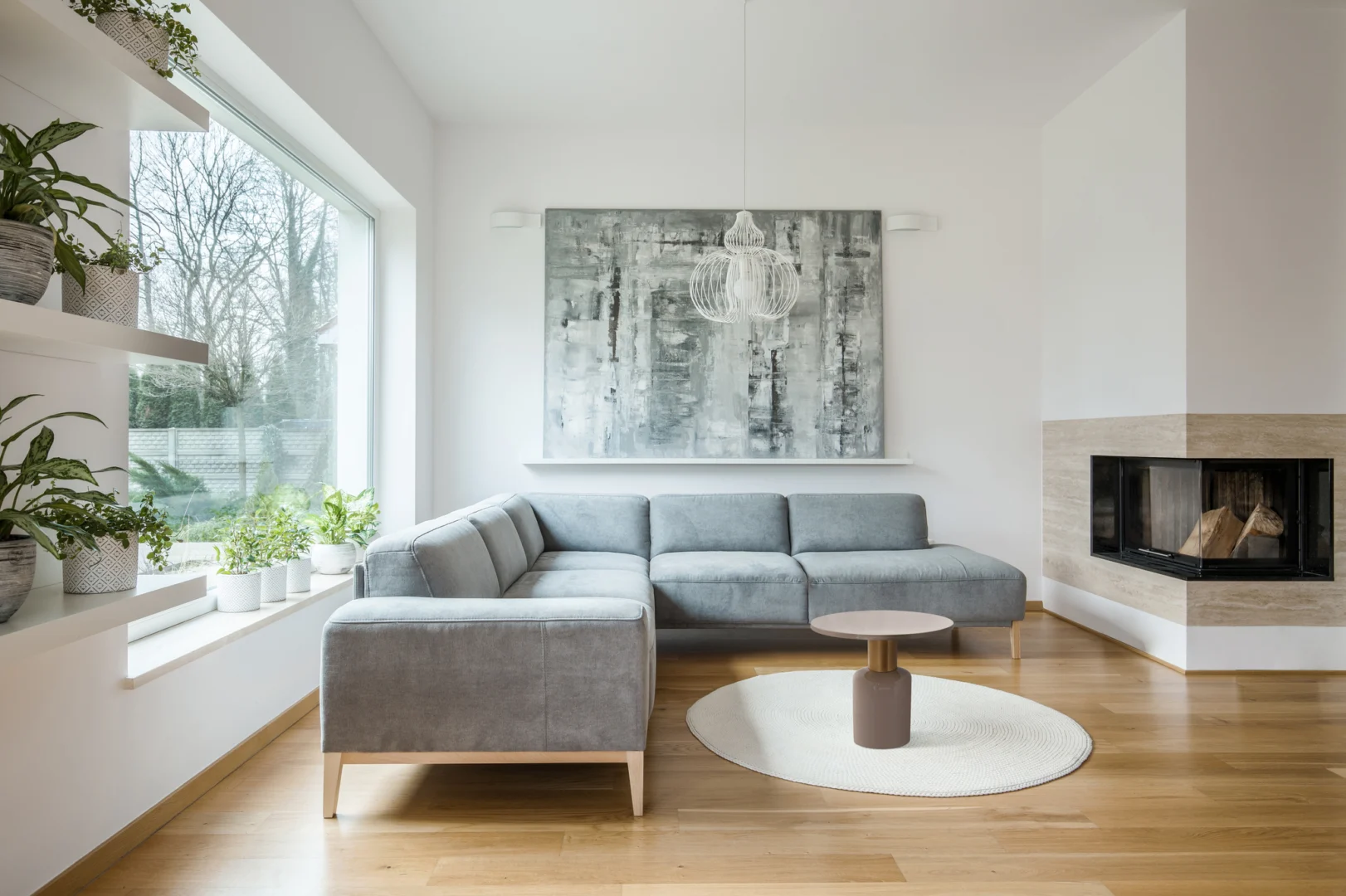 Een moderne woonkamer heeft een grijs bankstel, Bijzettafel Taupe Ijzer Design als salontafel, een cirkelvormig vloerkleed op de houten vloer, grote abstracte kunst boven de bank en planten in potten bij een groot raam en op wandplanken.