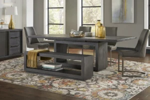 Een moderne eetkamer met de Rechthoekige eettafel Oxford in basalt grijs, vier grijze stoelen en een bijpassende bank met een mosterdkleurige deken. Op de bank staan decoratieve voorwerpen en boeken, terwijl grote ramen en een abstract vloerkleed de ruimte opfleuren.