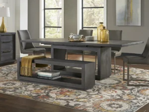 Een moderne eetkamer met de Rechthoekige eettafel Oxford in basalt grijs, vier grijze stoelen en een bijpassende bank met een mosterdkleurige deken. Op de bank staan decoratieve voorwerpen en boeken, terwijl grote ramen en een abstract vloerkleed de ruimte opfleuren.