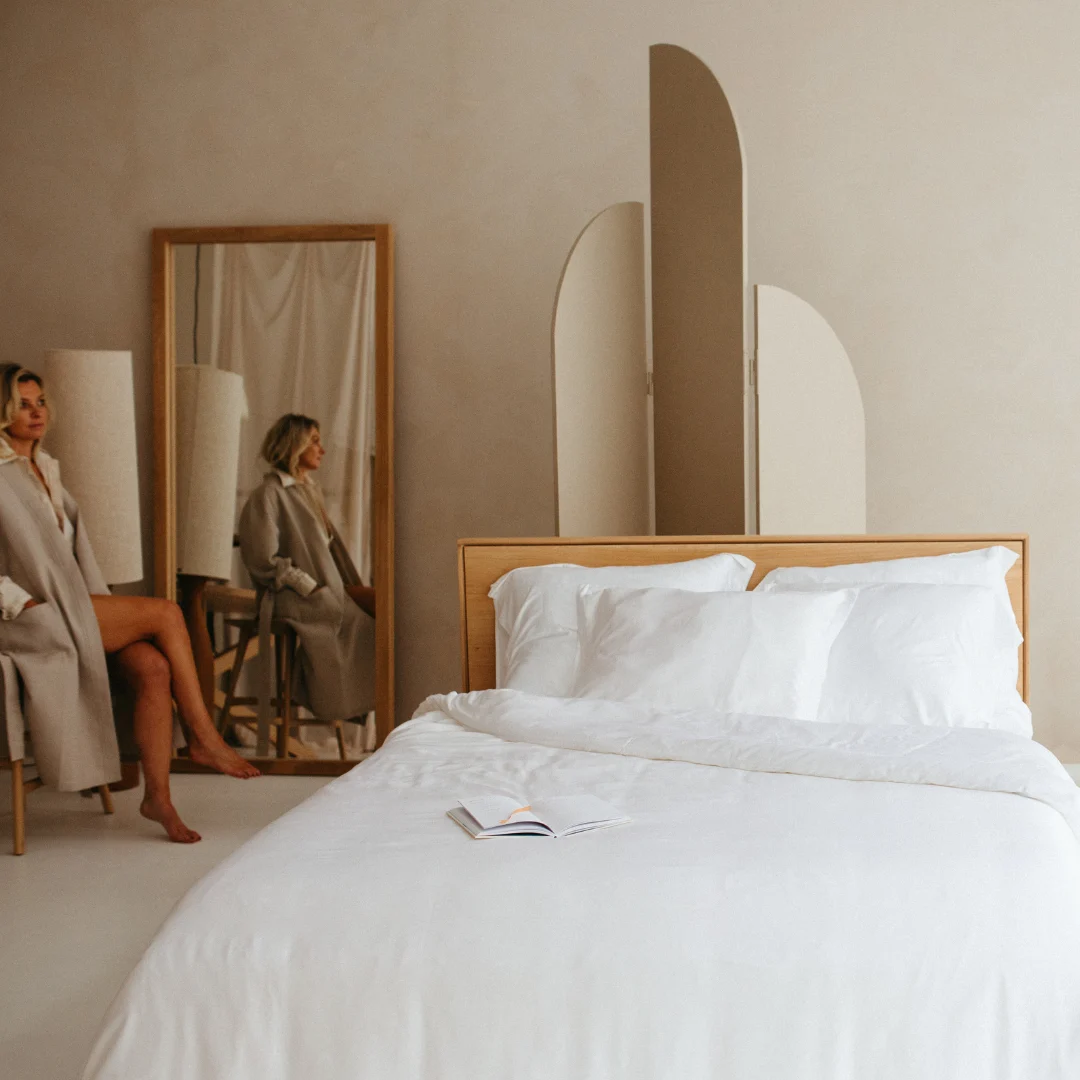 Een vrouw in een beige jas zit naast een grote spiegel in een minimale, moderne slaapkamer met lichte muren, abstract decor en een keurig opgemaakt bed bedekt met de Dekbedovertrek 140x220 Wit Snelle upgrade.