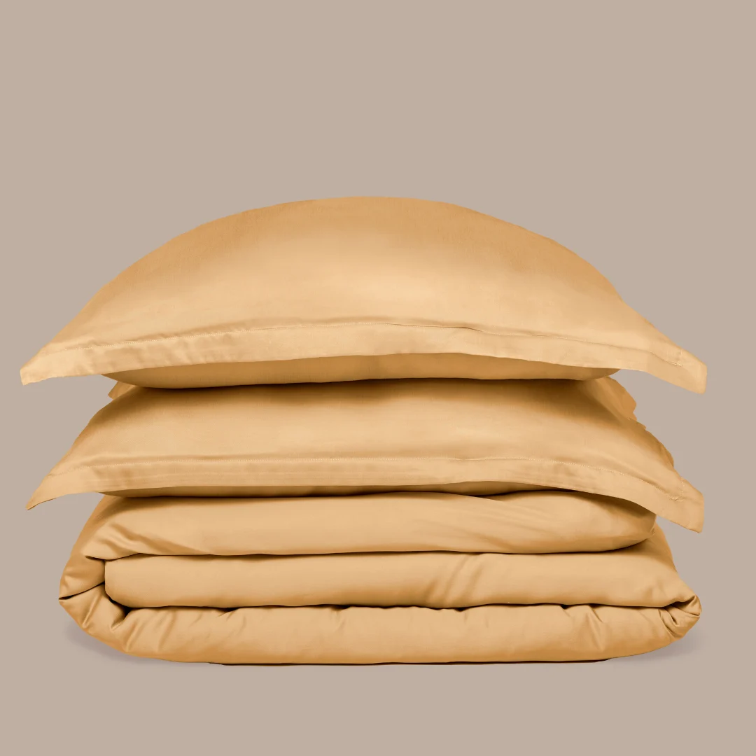 De Dekbedovertrek 200x200 Geel Luxe set, bestaande uit een dekbedovertrek en twee kussenslopen in goudbruin, is netjes gestapeld tegen een effen beige achtergrond.