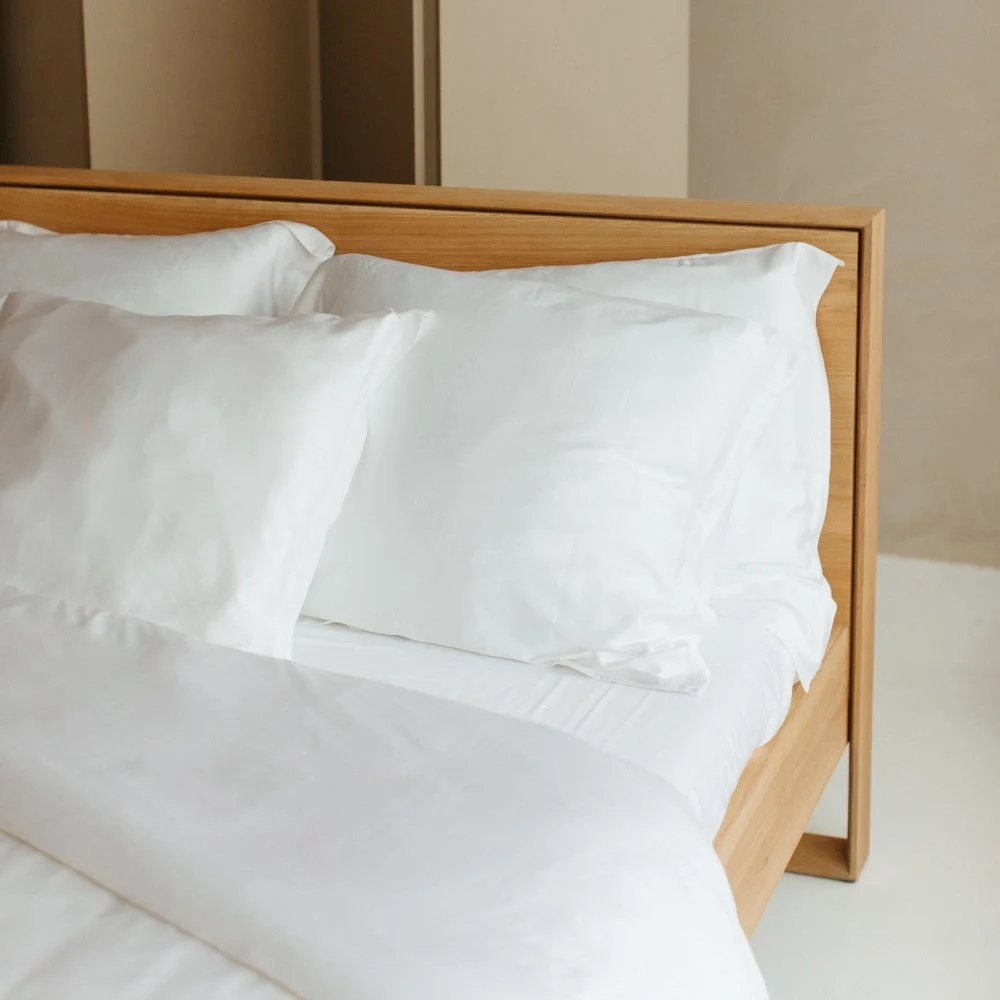 Detailopname van een bed met een houten frame en witte kussenslopen. Zachte belichting benadrukt het gladde, zijdezachte materiaal.
