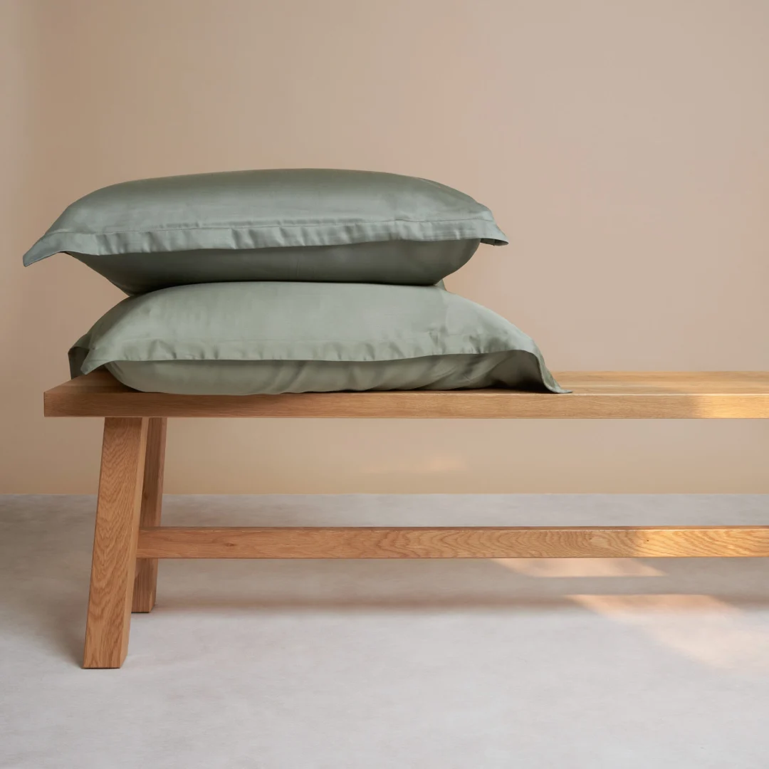 Twee groene kussenslopen van bamboe op een houten bankje, zachte belichting, beige achtergrond. De kussens zijn 65x65 cm groot.