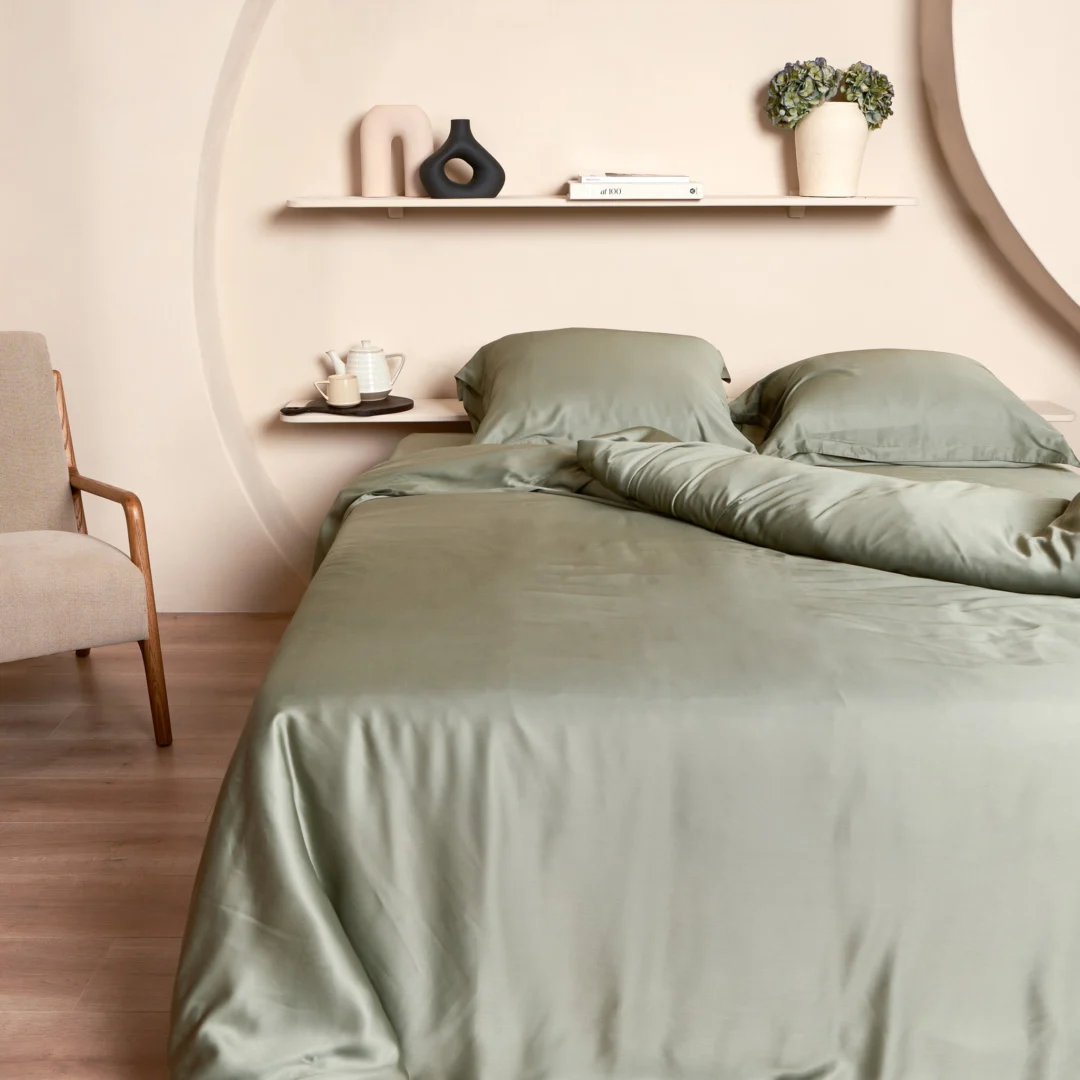 Slaapkamer met olijfgroen beddengoed, inclusief bamboe kussensloop 65x65. Zachte belichting, lichte muren, houten vloer en stoel.