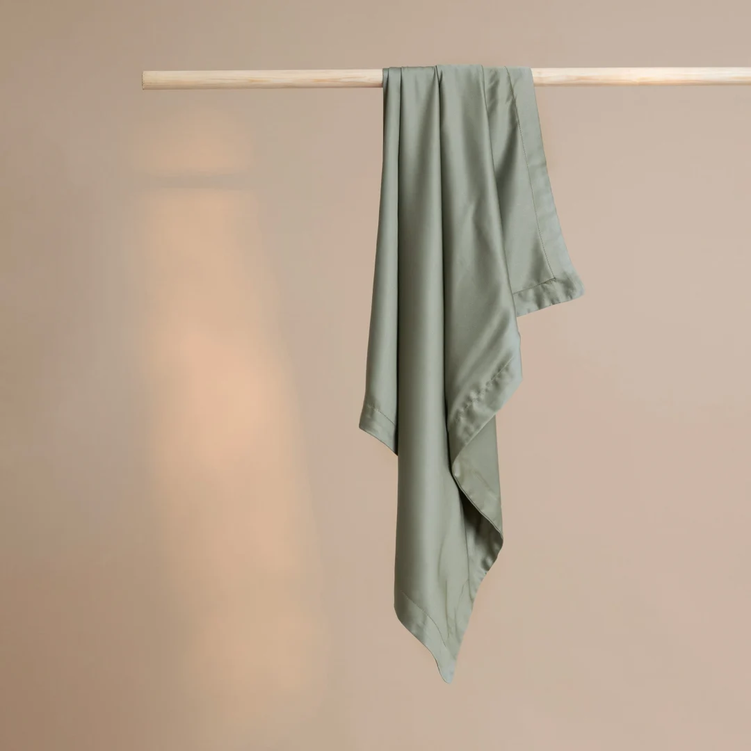 Zachte, groene bamboe kussensloop hangt aan een houten stok. Gladde stof met subtiele glans. Neutrale achtergrond. Minimalistisch en rustgevend.