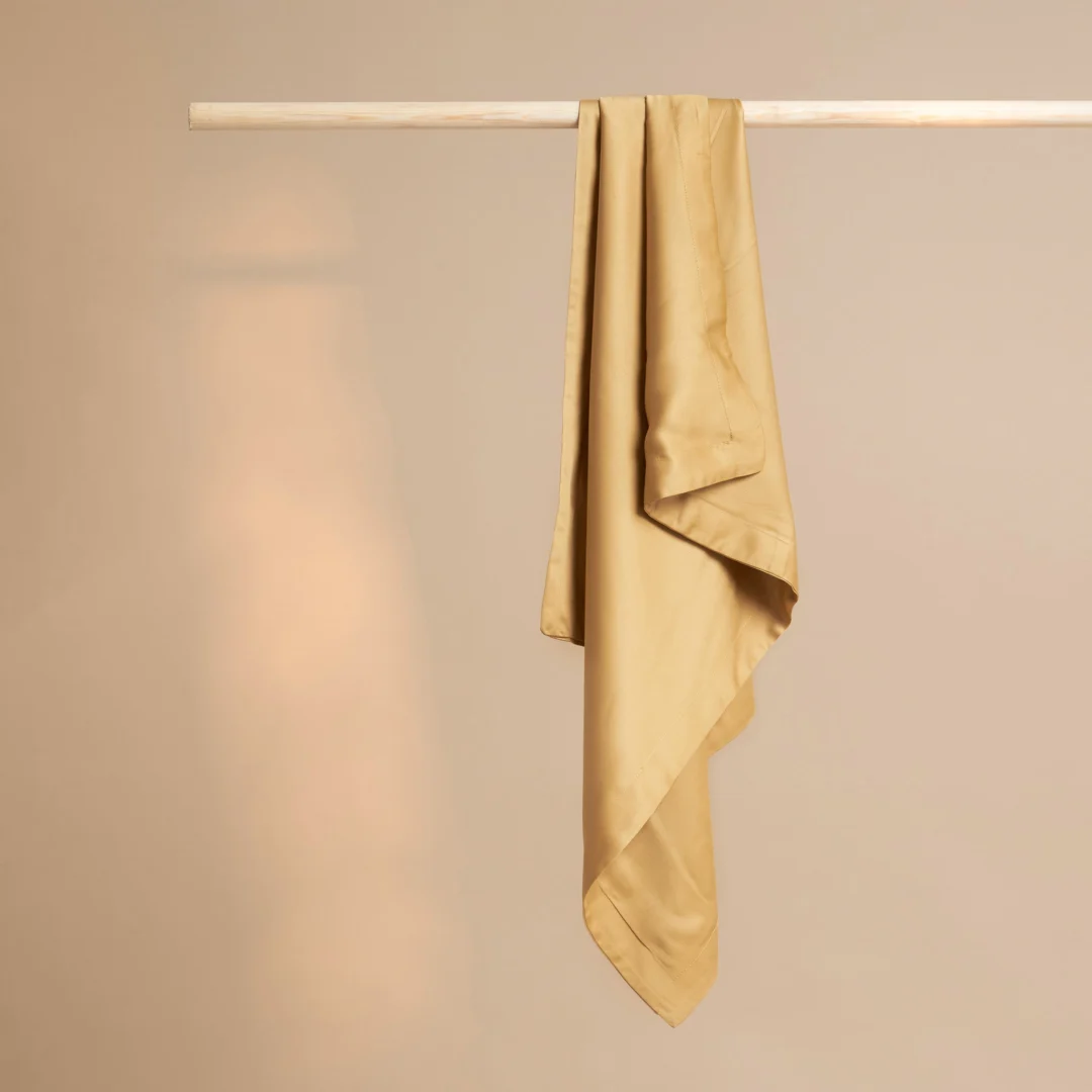 Gouden kussensloop van eco bamboe hangt aan een houten stang. Zachte belichting, effen achtergrond. Zijdezacht materiaal valt in plooien.