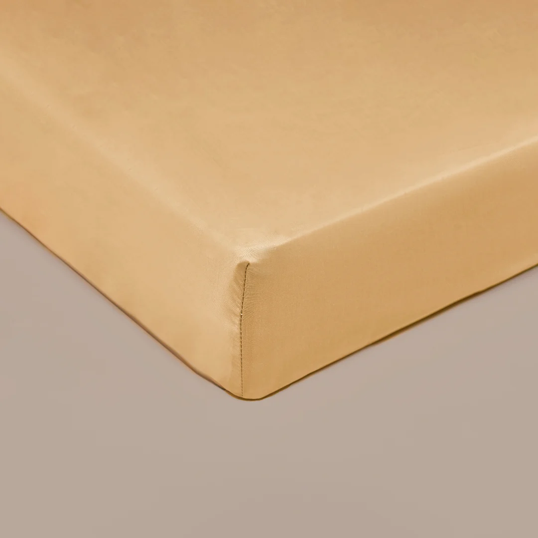 Detailopname van een okergeel hoeslaken, gladgestreken en strak gespannen. Zachte belichting accentueert de textuur. Rustige, neutrale achtergrond.