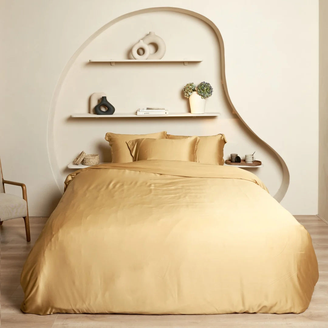 Okergeel hoeslaken van bamboe opgemaakt op een bed met kussens. Zachte belichting, lichte achtergrond met decoratieve planken.