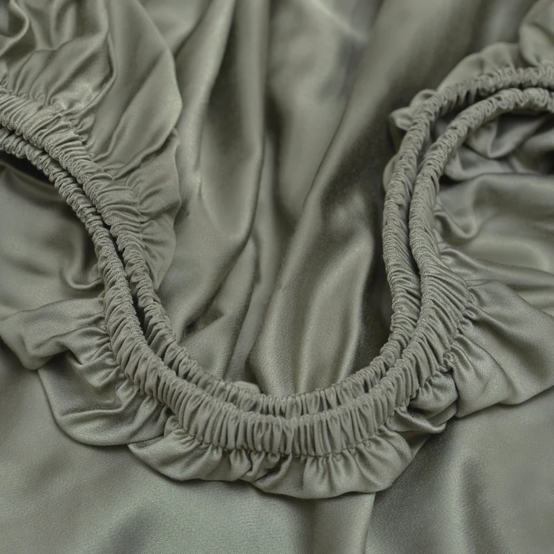 Close-up van olijfgroen hoeslaken met elastiek. Zachte stof met plooien, gladde textuur. Satijnen glans, warme belichting.