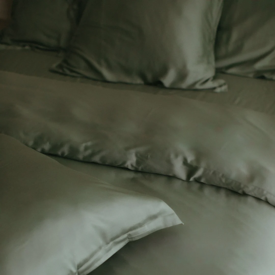 Close-up van een olijfgroen bamboe hoeslaken en kussenslopen. Zacht licht accentueert de gladde textuur van het beddengoed.